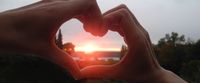 Hände zu Herz geformt (Herz-Symbol) vor Sonnenaufgang oder Sonnenuntergang. Die Sonne scheint direkt durch das geformte Herz.